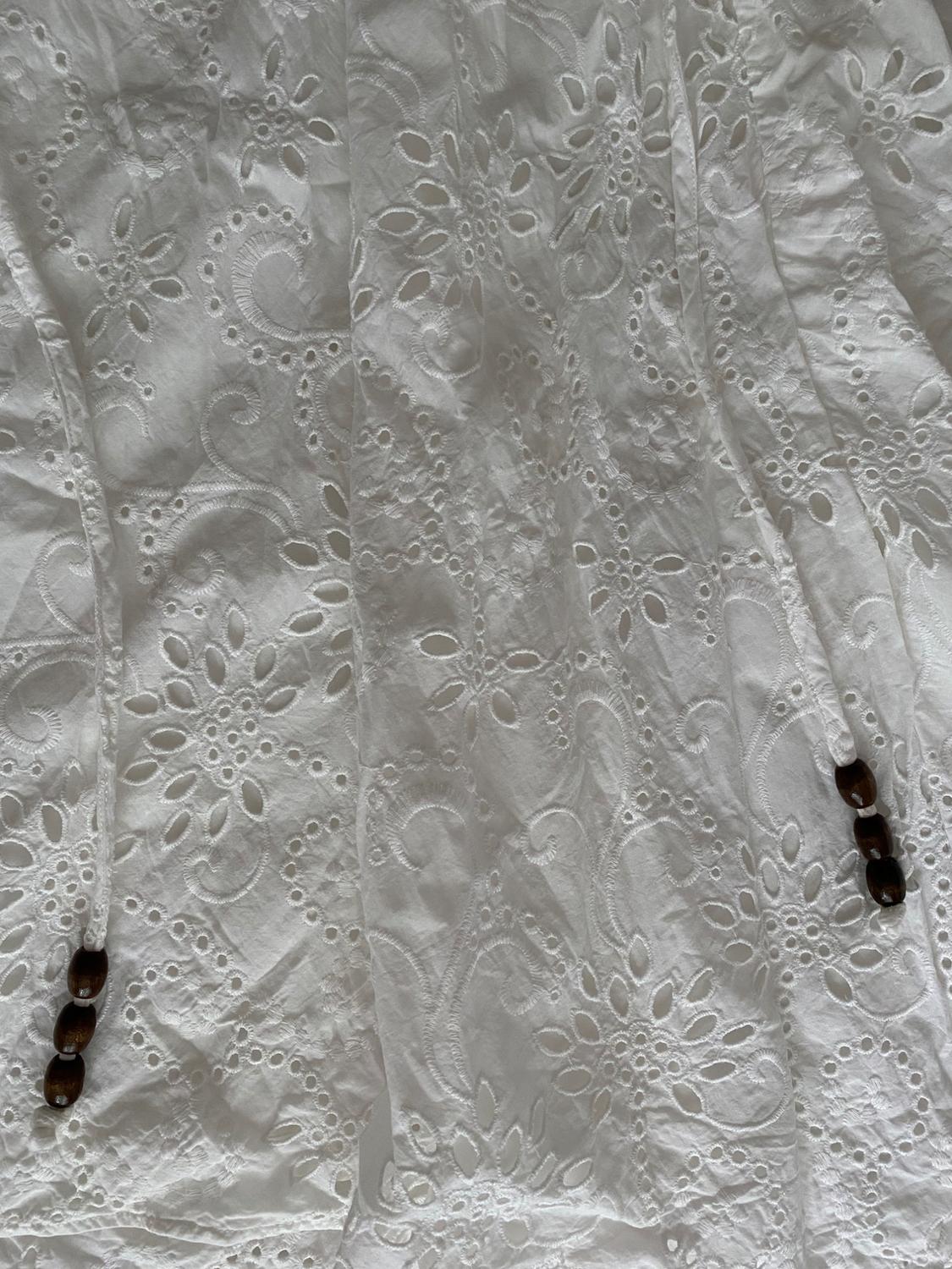 Close-up of white fabric with perforated floral embroidery and thin laces finished with dark wooden beads.