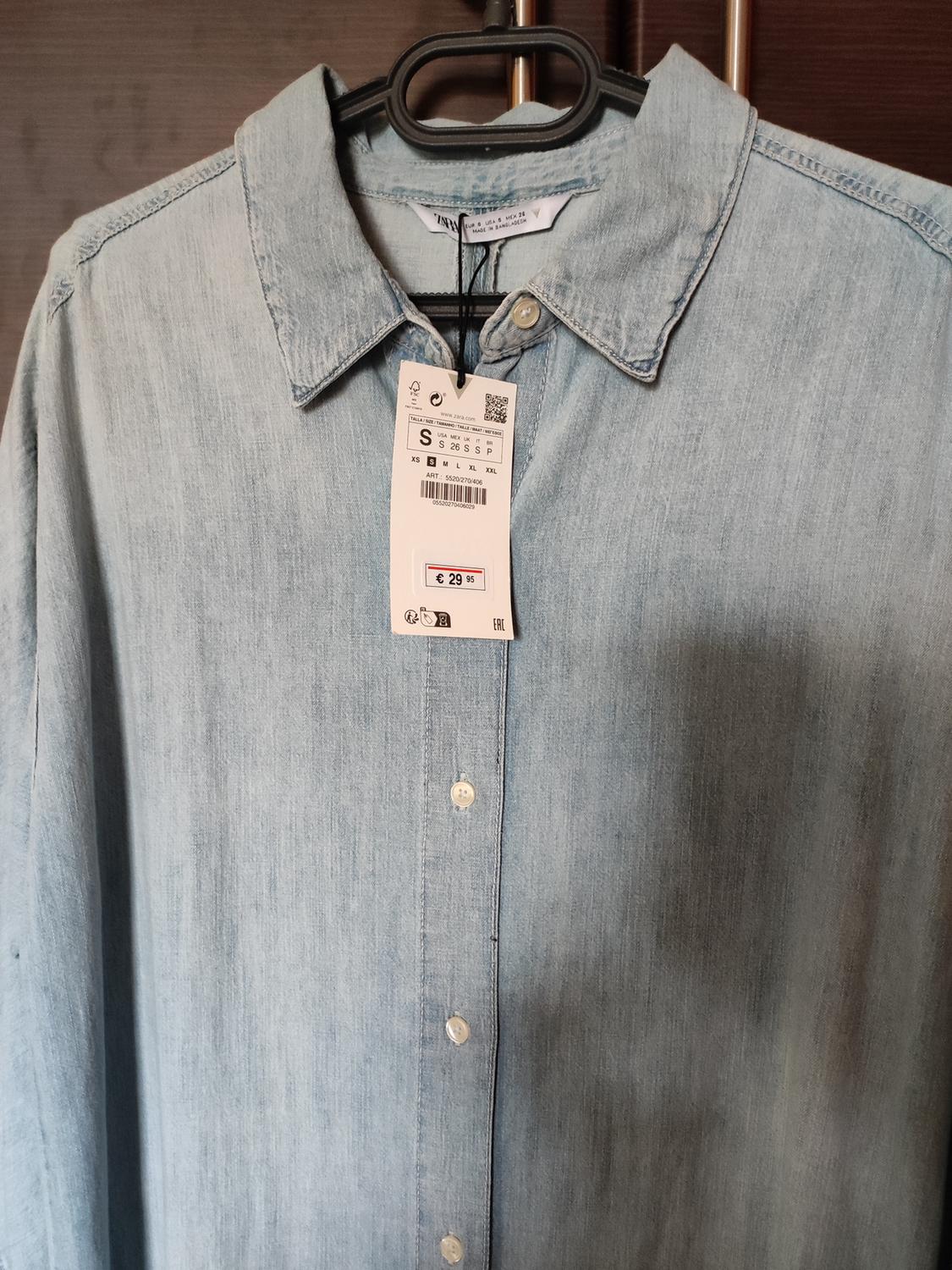 Light blue denim shirt hanging on a hanger, with a price tag of €29.95 and size S visible. Dark wooden background.