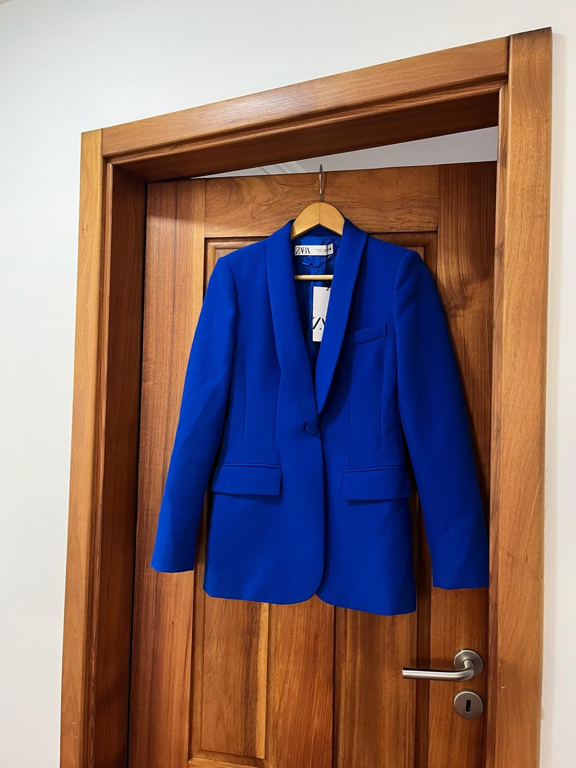 Bright blue blazer hanging on a hanger over a wooden door. The text ZARA can be read on the label.