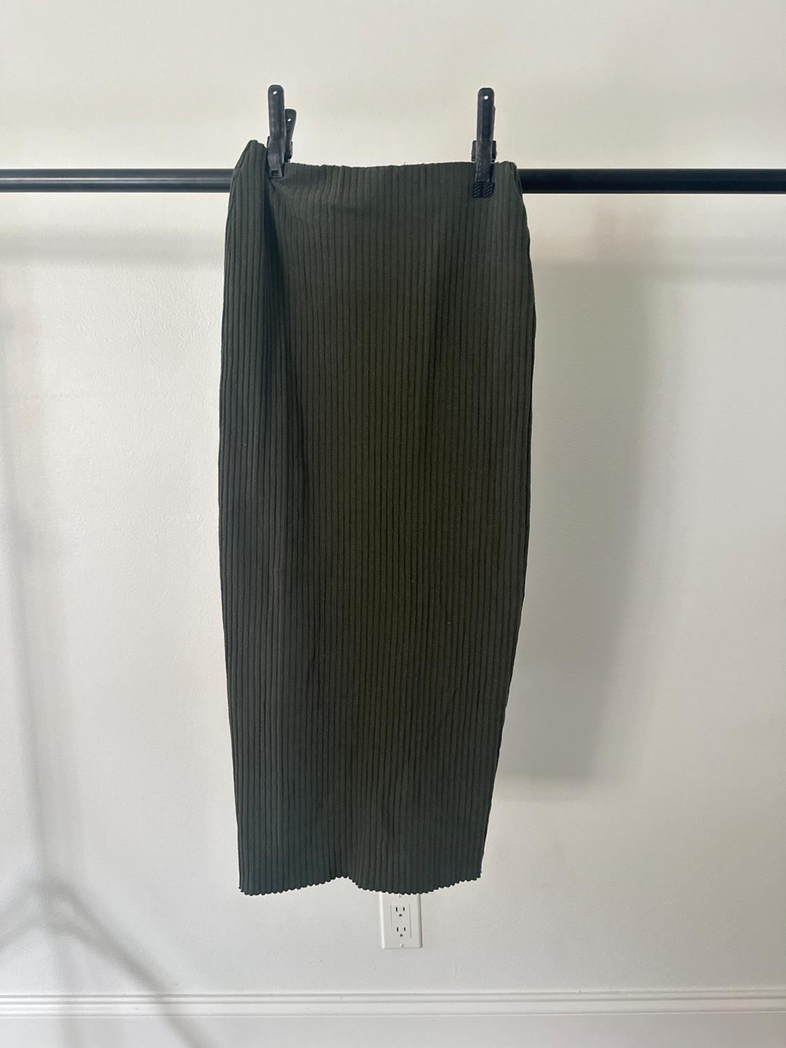 A long, dark green skirt with a ribbed texture, hanging on a black rail against a white wall.