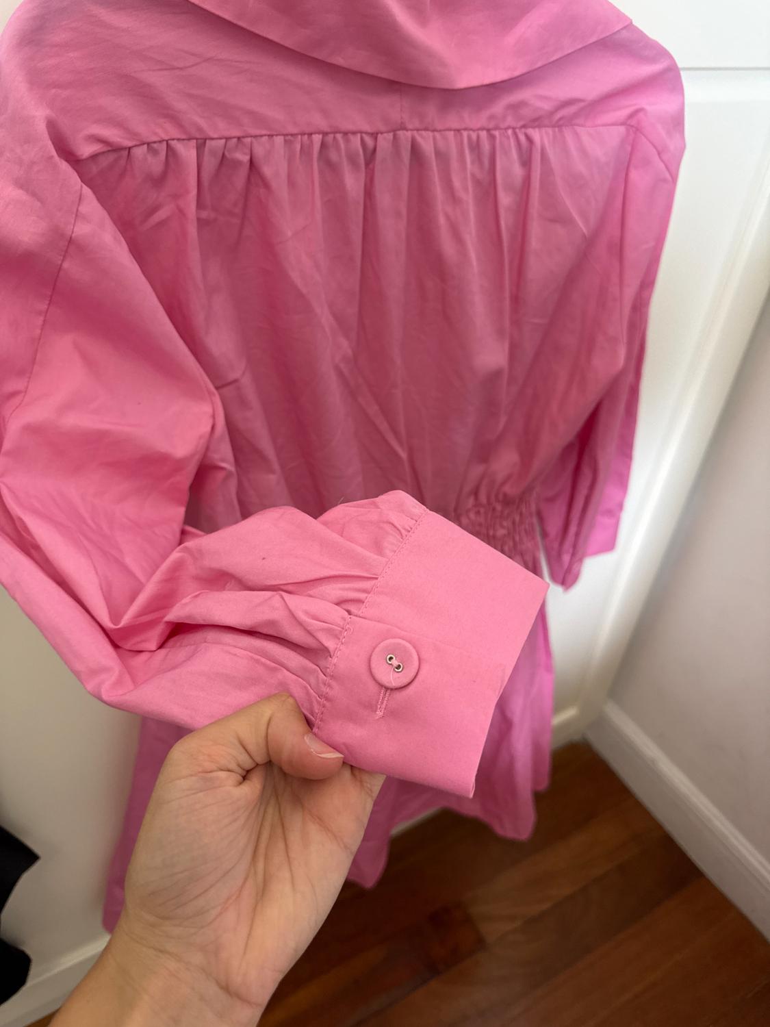 Hand holding the buttoned cuff of a pink garment. The pleated back, elasticated waist, white door, and wooden floor are visible.