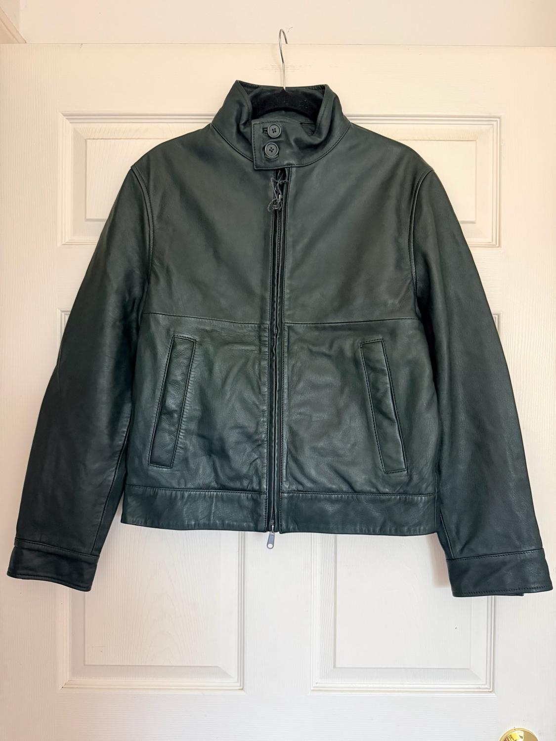 Black leather jacket with a zipper, high collar with buttons, and side pockets, hanging on a hanger over a white door.