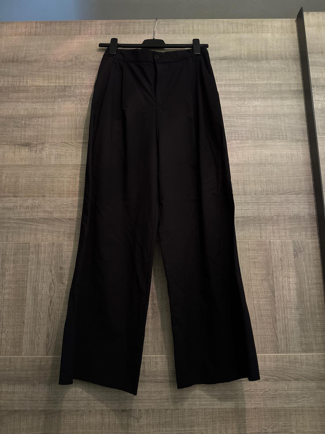 Wide-leg black trousers with pleats and a raw hem, hanging on a black hanger in front of a wooden wardrobe.