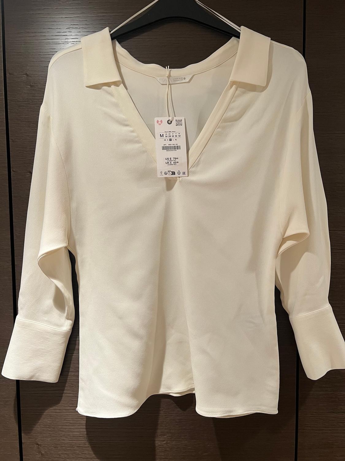 White long-sleeved open-collar blouse, hanging on a black hanger. Visible tag with size and price details.