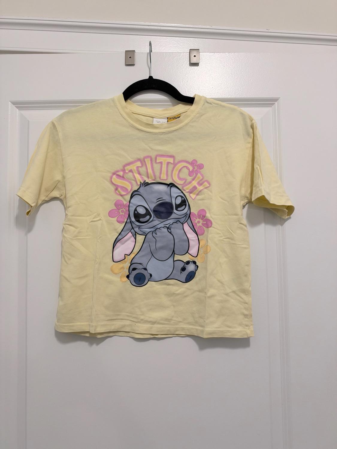 Yellow short-sleeved t-shirt hanging on a white door. Front print of Stitch with flowers and the text STITCH.