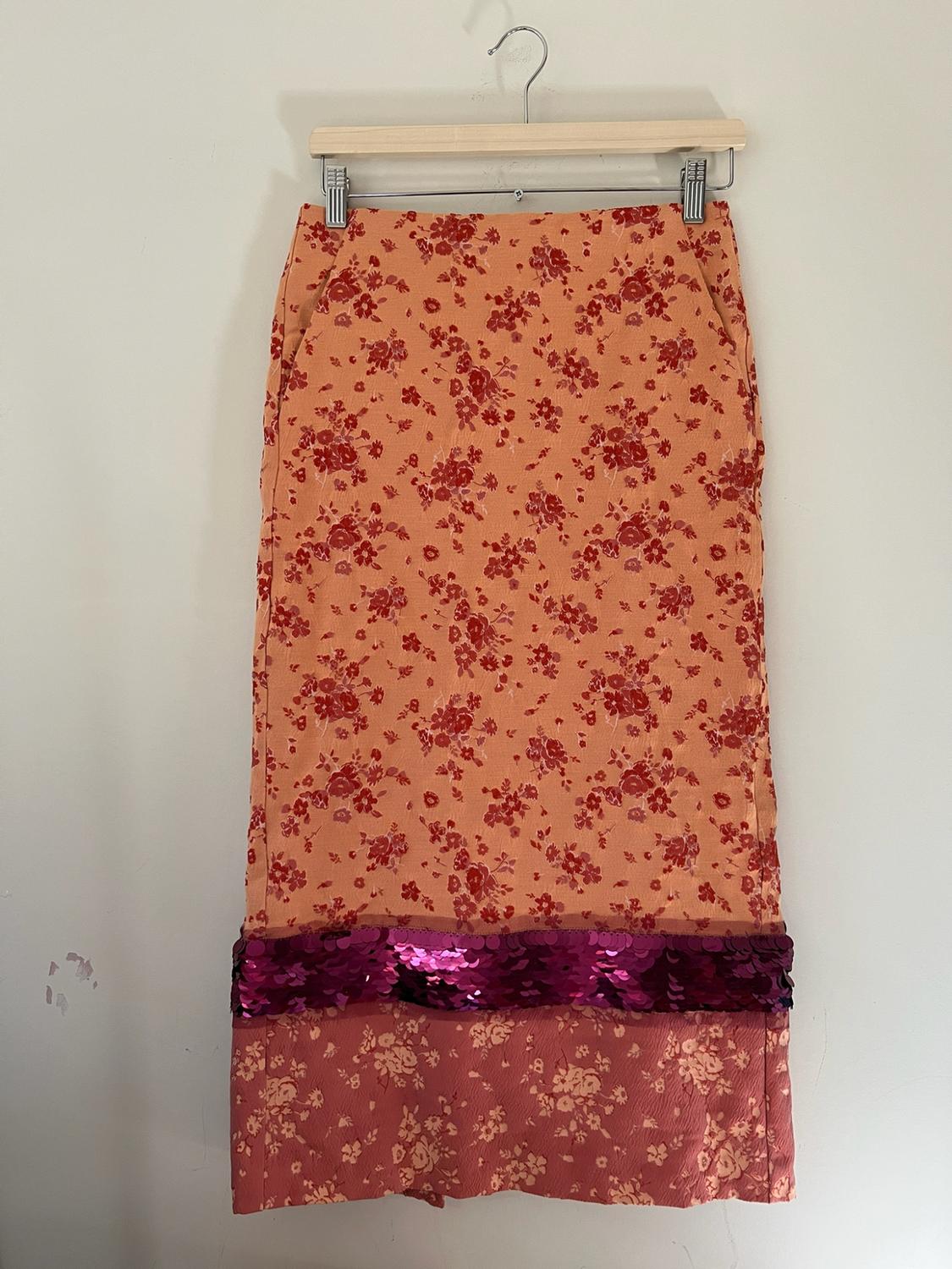 Long floral skirt hanging on a hanger against a white background. Design in orange and pink tones with a purple sequin trim at the hem.