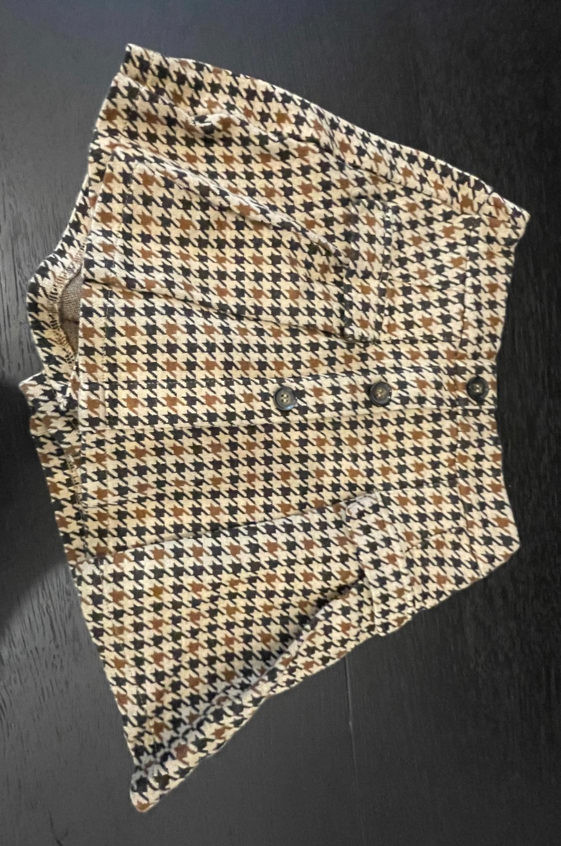 Short skirt with a black, white and brown houndstooth pattern, featuring three front buttons, on a dark surface.