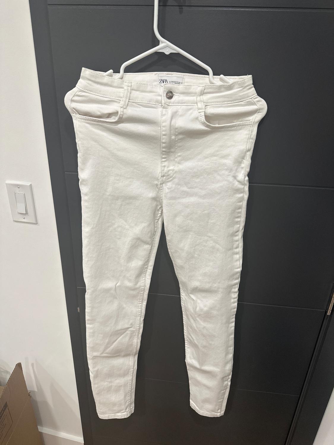 White pants hanging on a hanger in front of a dark door. Visible tag on the waistband with the text "ZARA WOMAN".