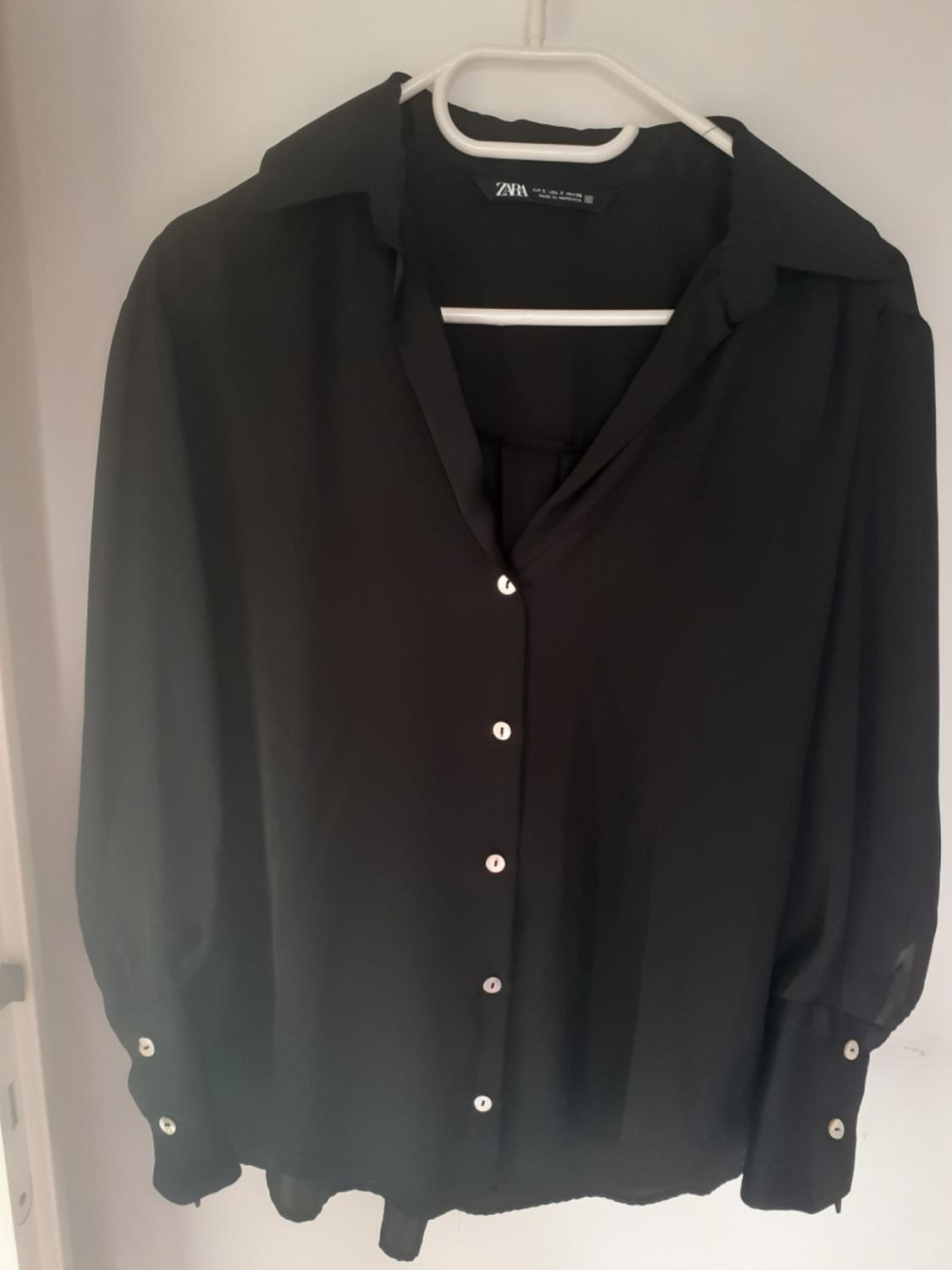 Black long-sleeved shirt with white buttons, hanging on a white hanger. The neck label reads ZARA.