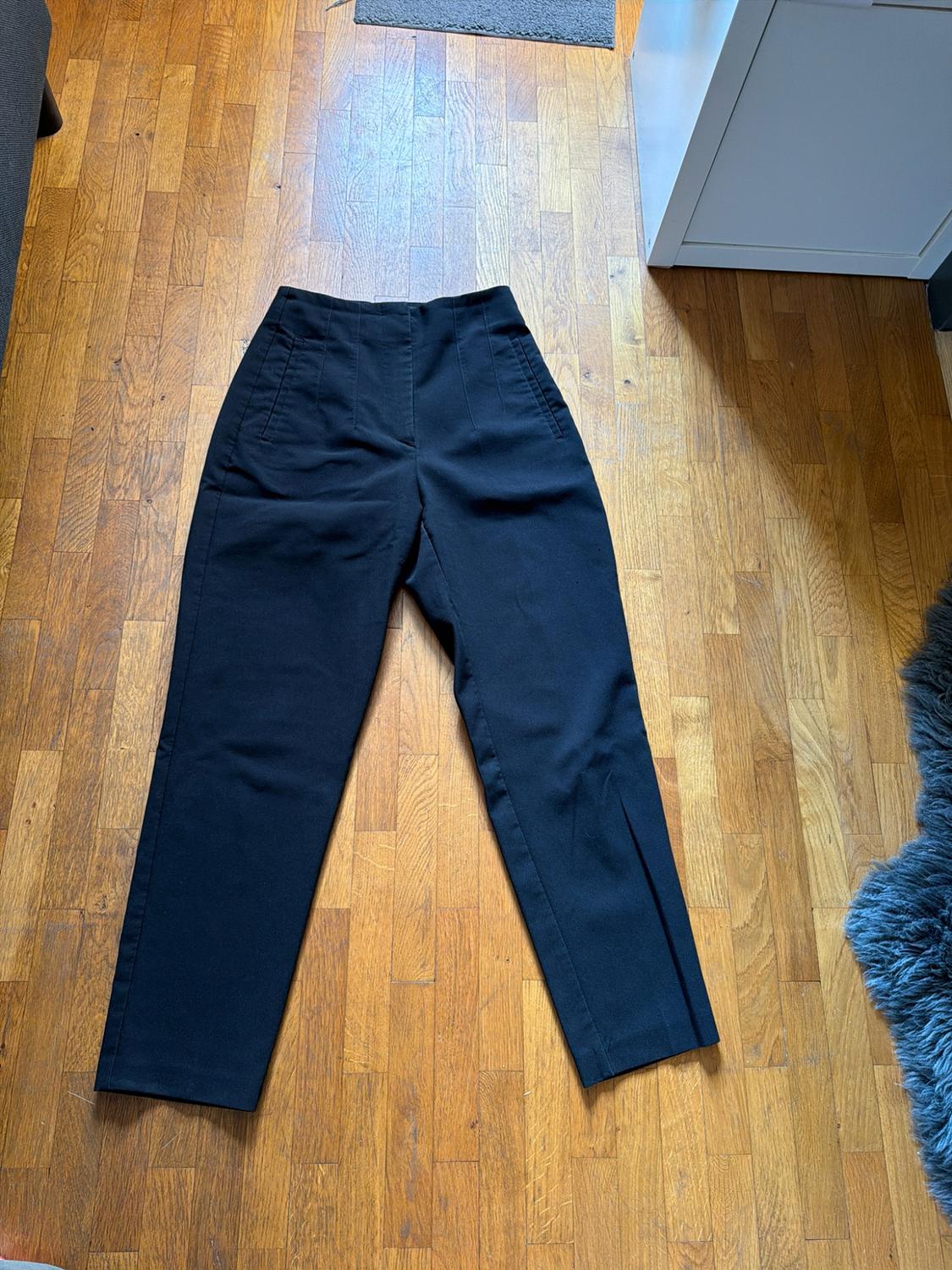 High-waisted black trousers with pleats, laid out on a wooden floor next to a white piece of furniture and a grey rug.