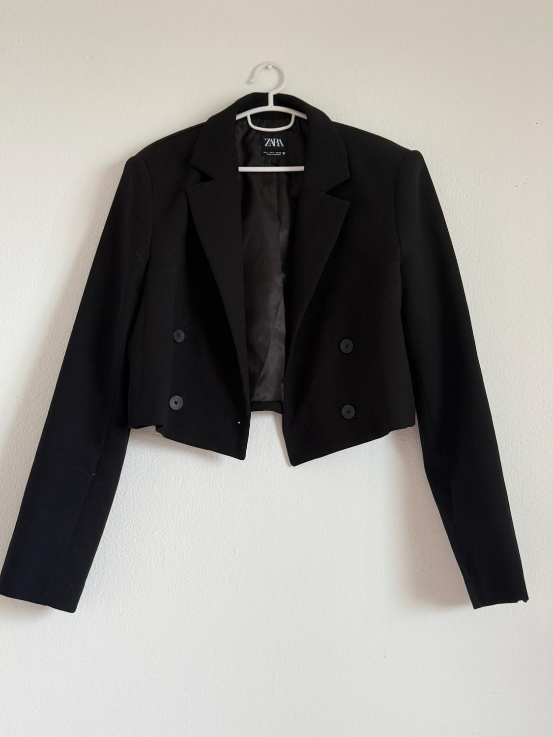 Black double-breasted cropped blazer with buttons, hanging on a white hanger against a white wall. Label with the text ZARA.