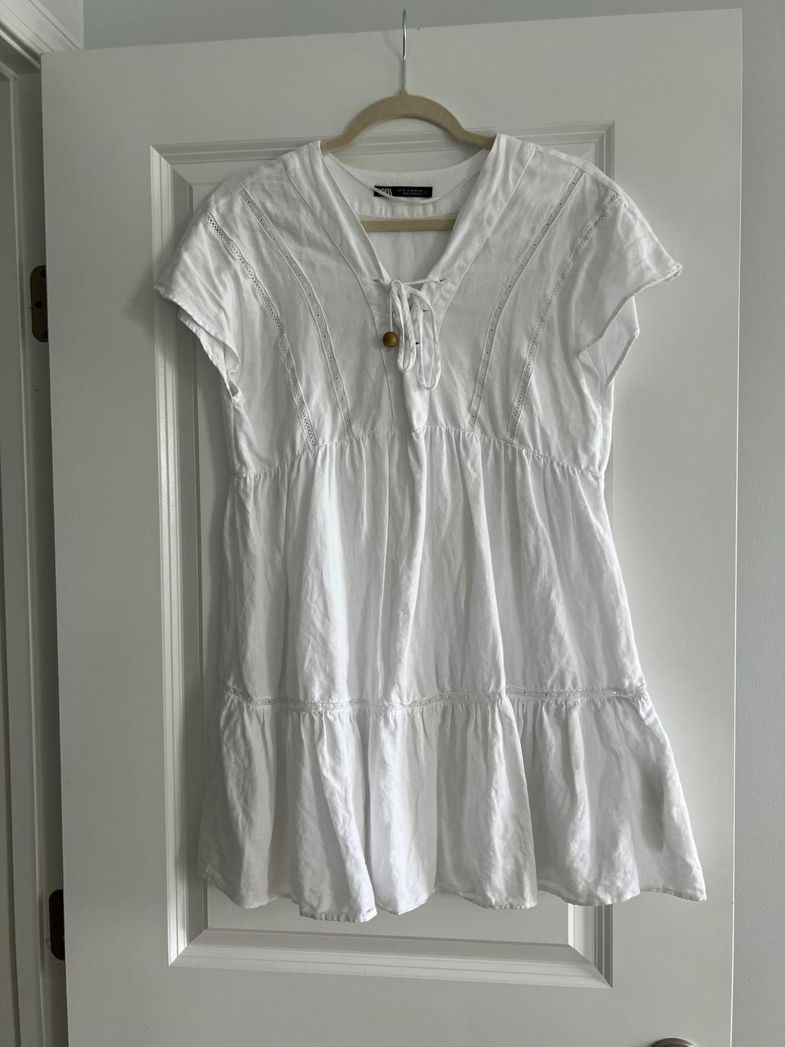 White short-sleeved dress hanging on a hanger over a white door. Lace and cord details on the neck.