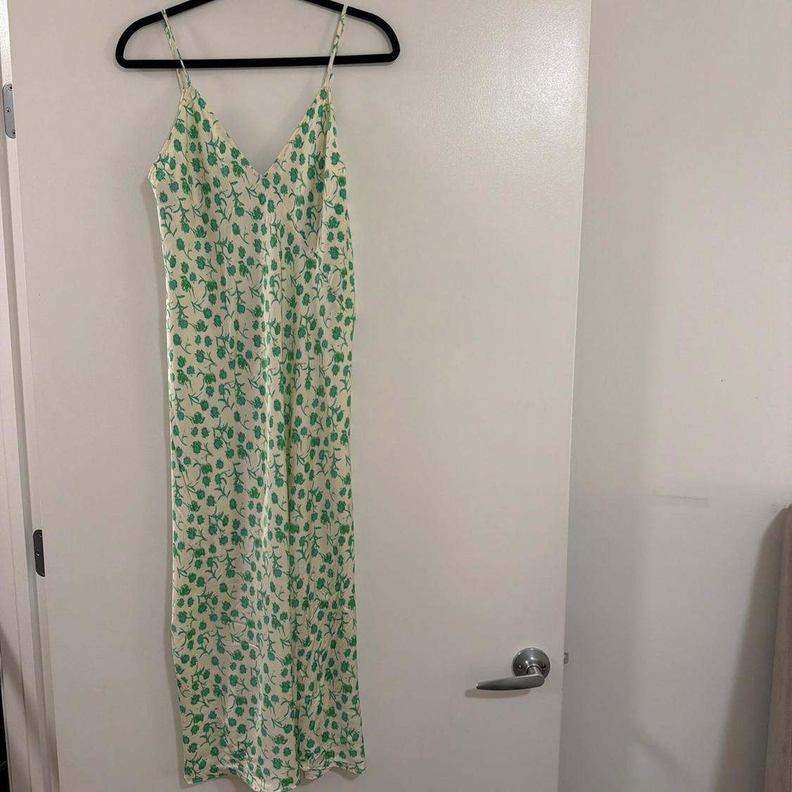White slip dress with a green floral print and spaghetti straps, hanging on a black hanger on a white door.