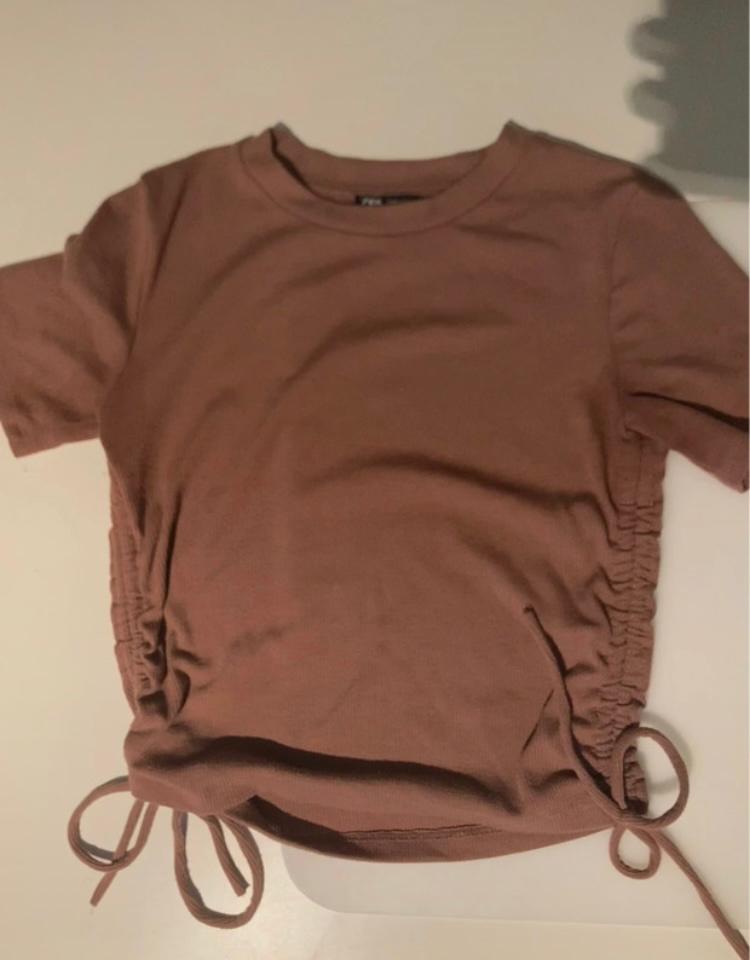 Brown short-sleeved T-shirt with side ruching and ties at the bottom, on a white surface.