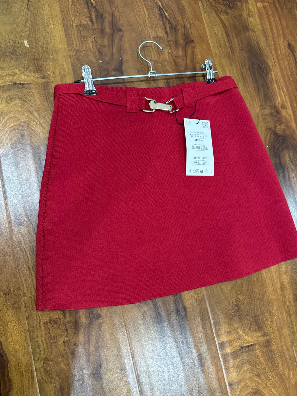 Red skirt with gold belt hanging on a hanger over a wooden floor. Label visible with price in dollars and pounds.