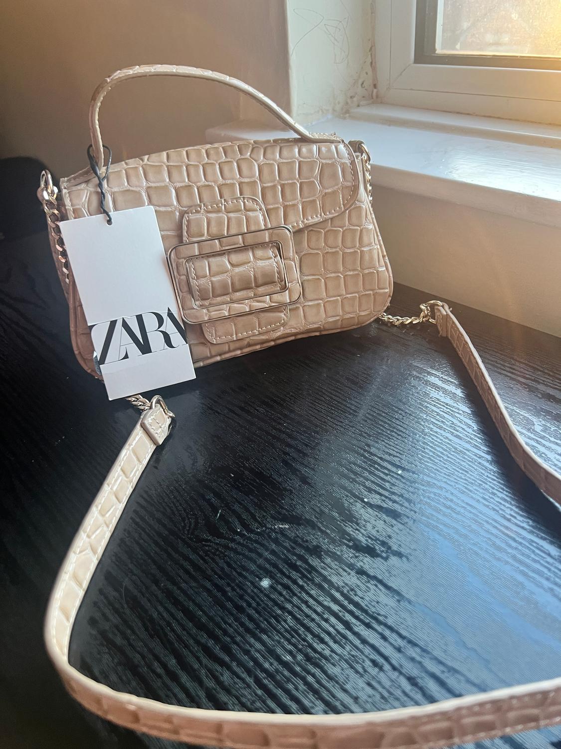 Beige crocodile-texture handbag on black table, "ZARA" tag hanging, natural light coming through the window.