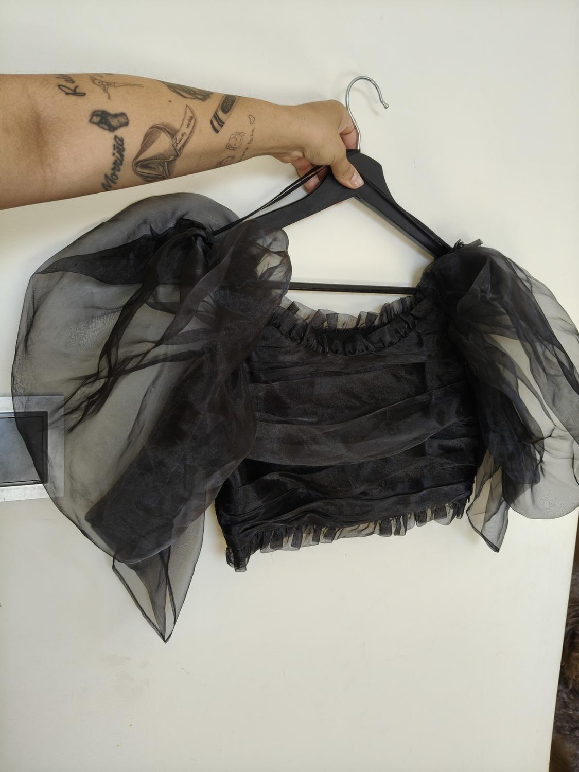 Tattooed arm holding a hanger with a translucent black top with a ruched bodice and puff sleeves against a white background.