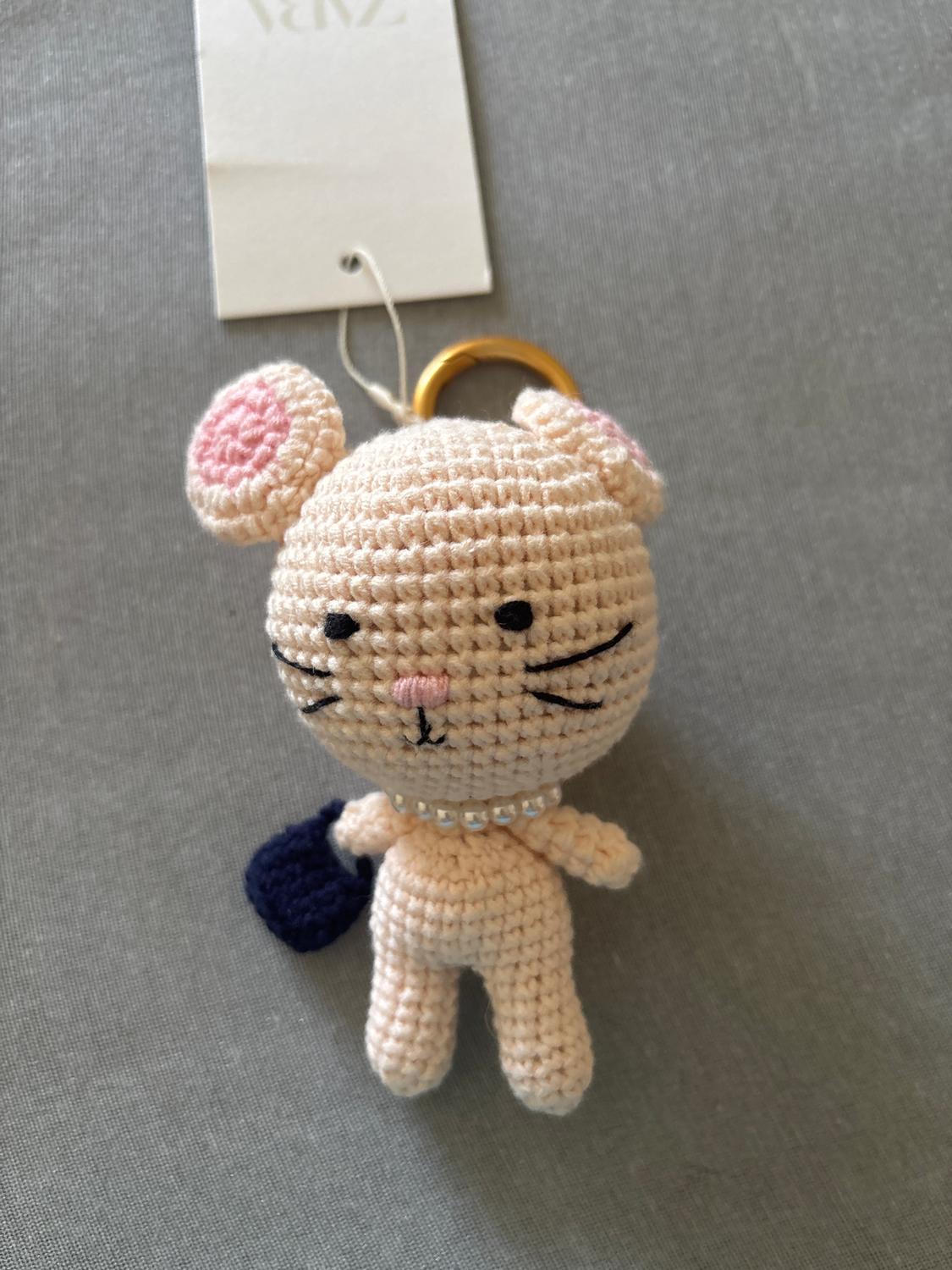 Beige crochet cat keyring, pink ears, pearl collar, and blue bag. Grey background, white tag at the top.
