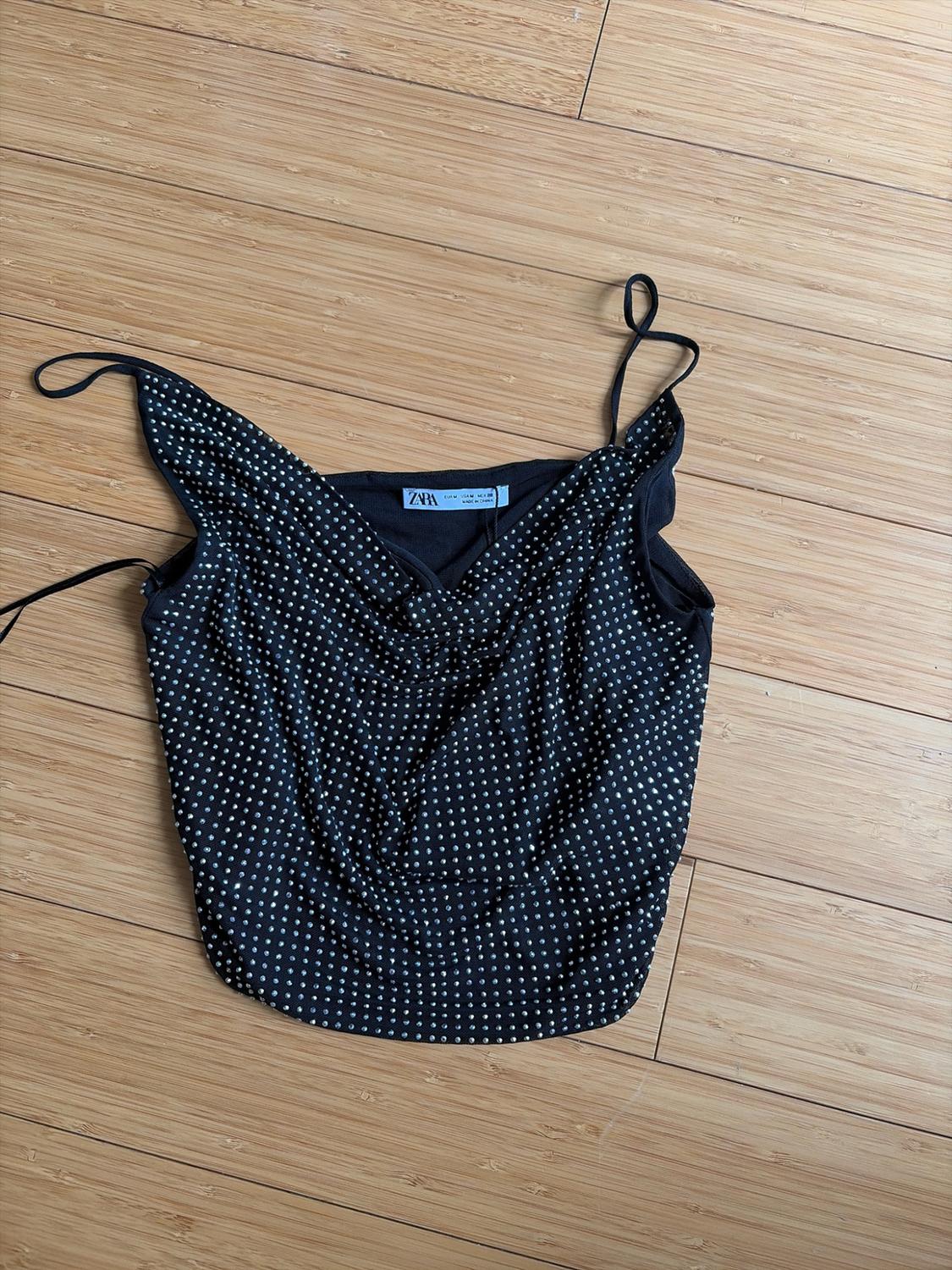 Black thin-strap camisole top with silver appliqués and a draped neckline, on a wooden floor. ZARA label.
