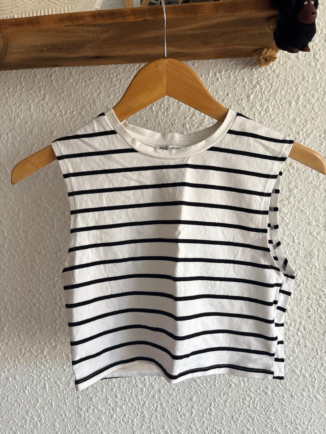 White sleeveless cropped top with black stripes hanging on a wooden hanger in front of a white wall. ZARA label visible.