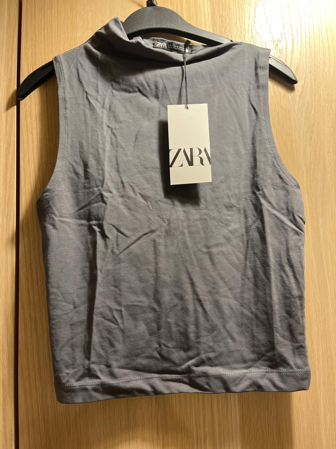Grey sleeveless top hanging on a black hanger, with visible Zara tag, on a light wooden background.