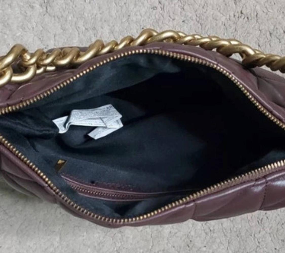 Open dark brown quilted bag, showing its black interior with white labels, zip and a thick gold chain.
