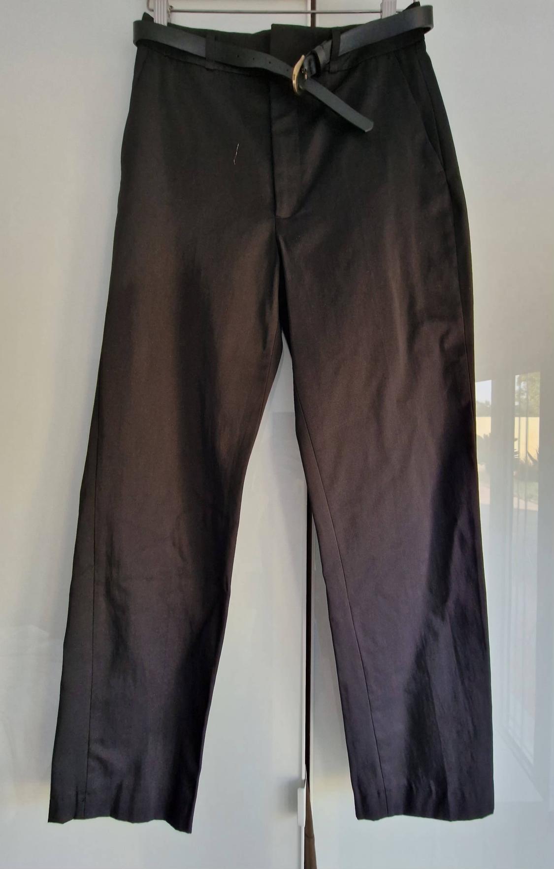 Black trousers with a matching belt and gold buckle, hanging on a hanger against a bright white background with light reflections.