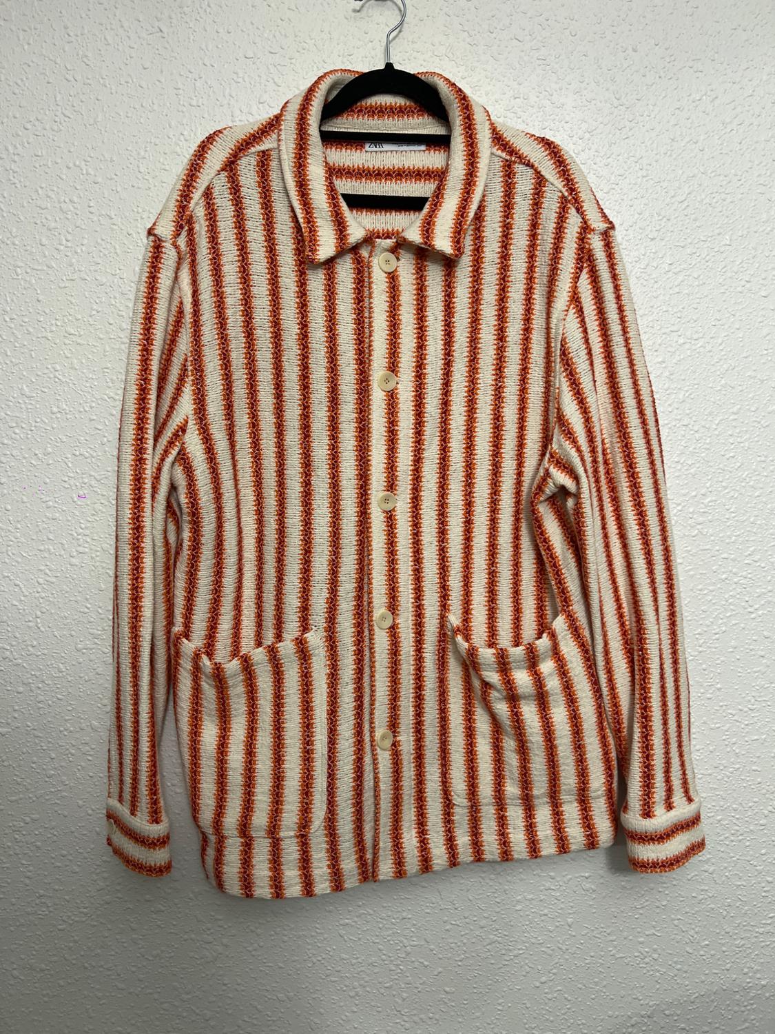 Knitted jacket with red and white vertical stripes, front buttons and large pockets, hanging on a black hanger.