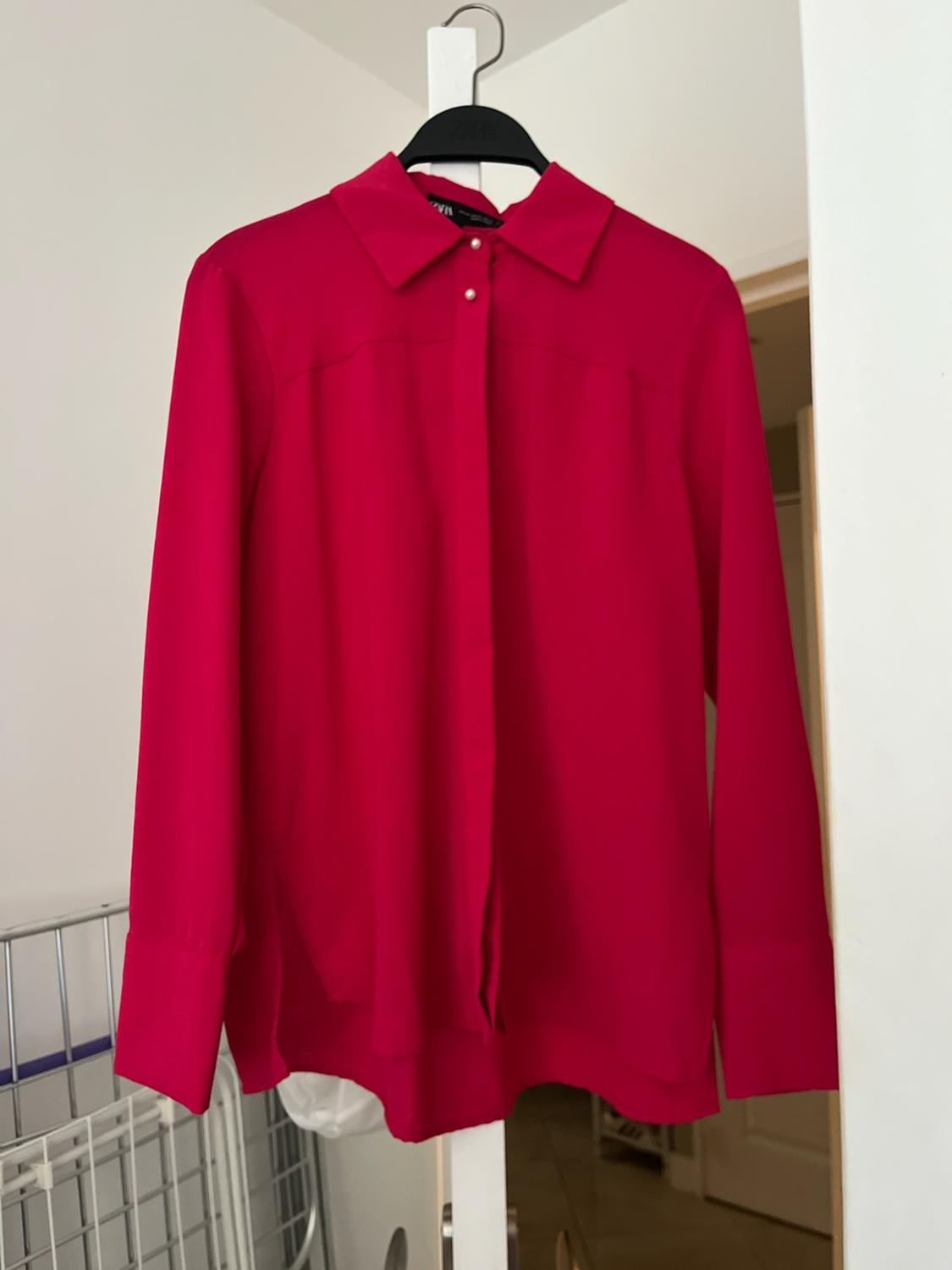 ZARA fuchsia long-sleeved shirt with a collar, pearlescent buttons and a concealed placket, hanging on a black hanger inside a house.