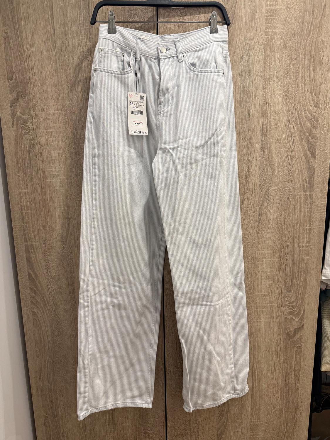 White and grey striped trousers hanging on a hanger against a wooden background. Visible label with barcode.