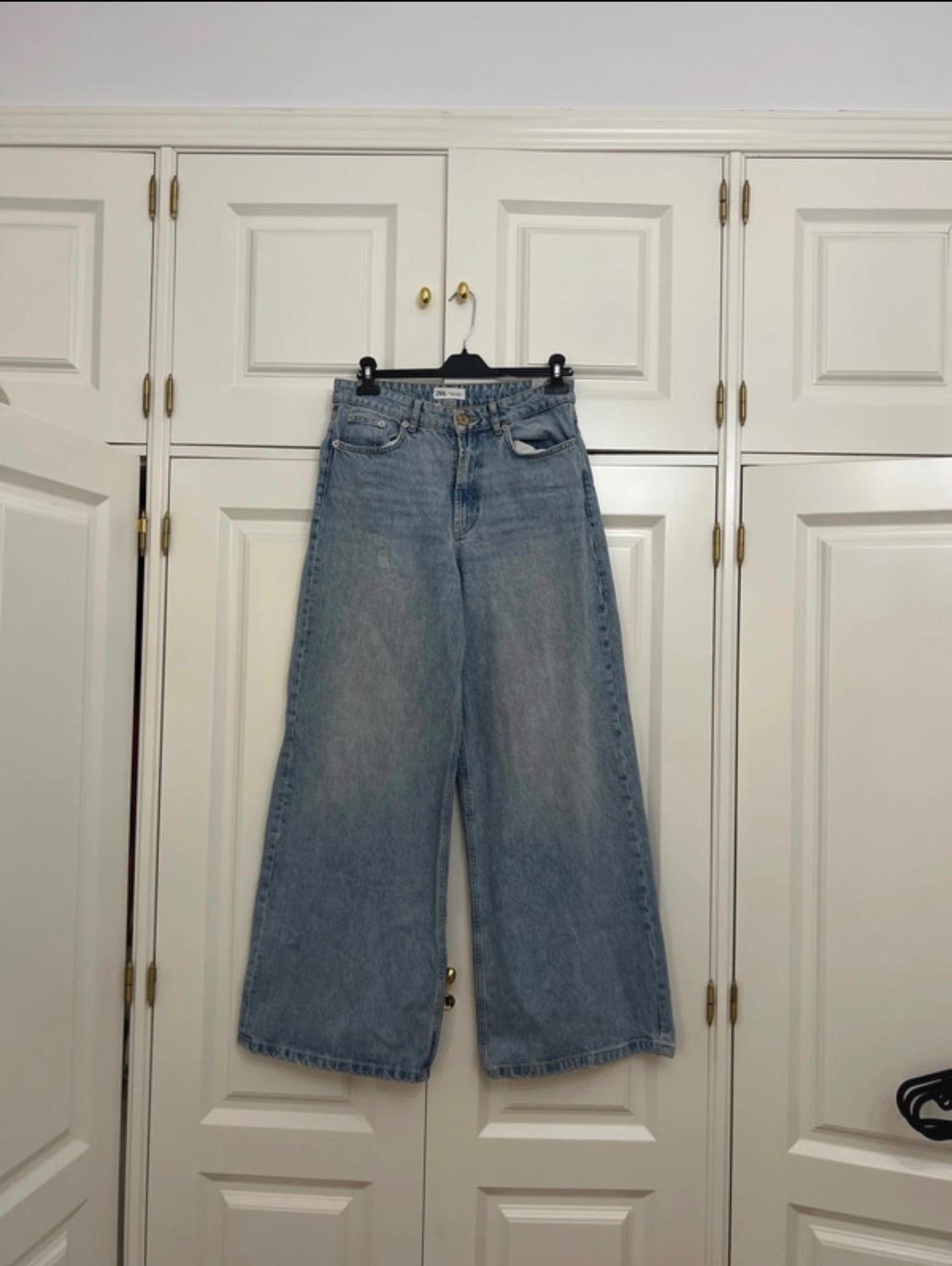 Light blue wide-leg jeans hanging on a black hanger in front of a white wooden wardrobe with gold handles. Casual style.