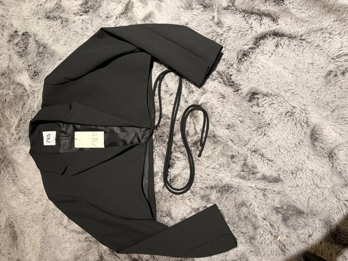 Short black jacket with a belt on a fluffy gray rug. Visible tag with the "ZARA" logo.
