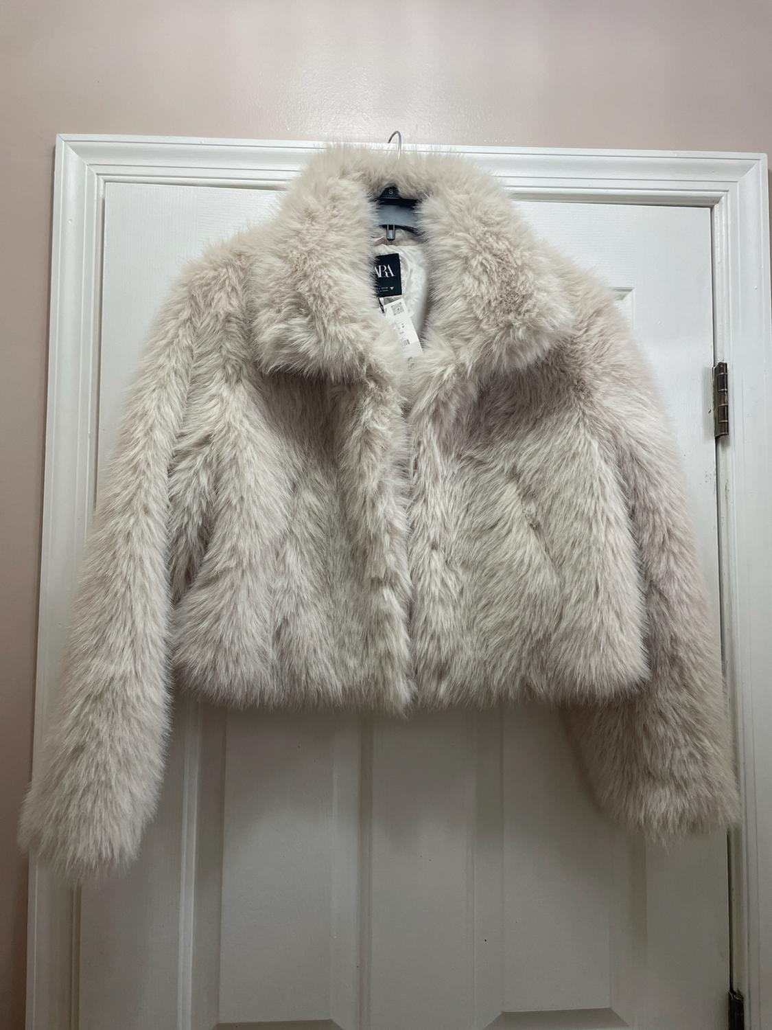 Cream-colored faux-fur jacket hanging on a white door. The black label with the text ZARA is visible at the collar.