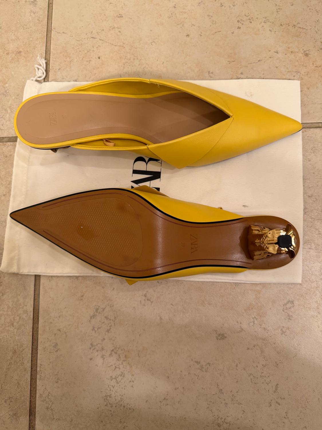 Sharp-toed yellow shoes on a white bag with "ZARA" text. Brown sole with gold detail on the heel.
