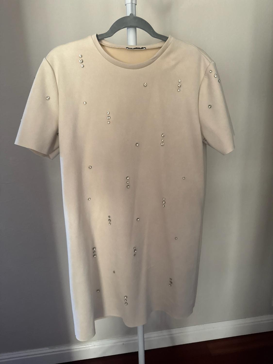 Short beige short-sleeve dress with scattered shiny rhinestone details, hanging on a gray hanger in front of a light-colored wall.