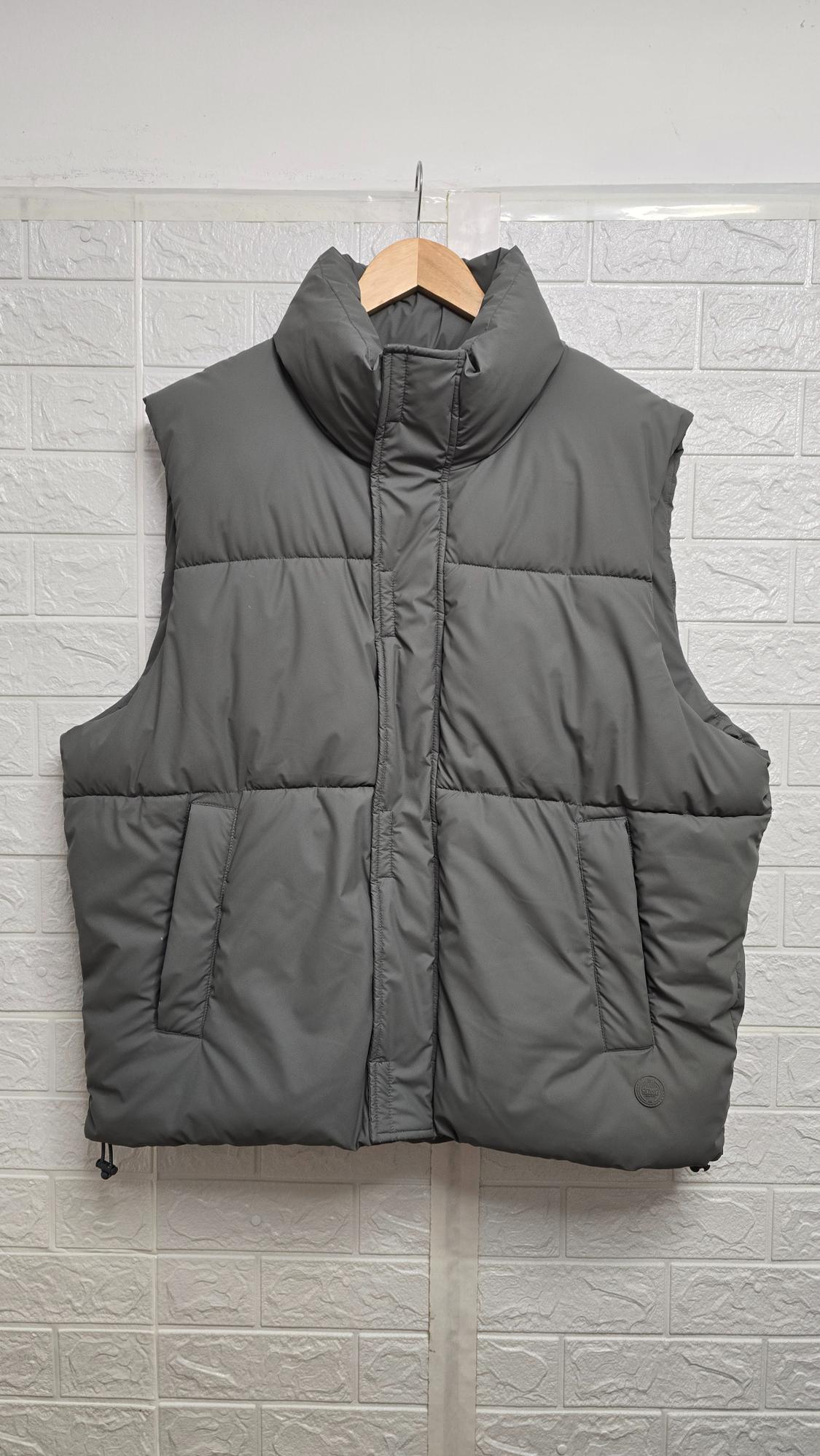 Grey padded gilet hanging on a wooden coat hanger, white brick-textured wall background.