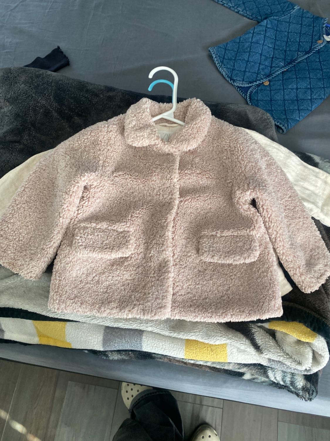 Light pink wool coat with pockets, hanging on a hanger over a bed with colourful blankets. Background with blue jacket.