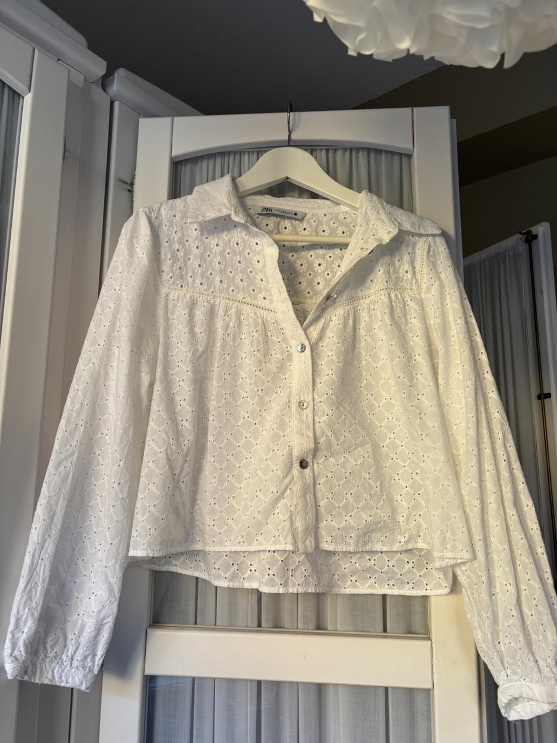 Long-sleeved white blouse with broderie anglaise, classic collar and buttons down the front, hanging on a hanger in front of white doors.