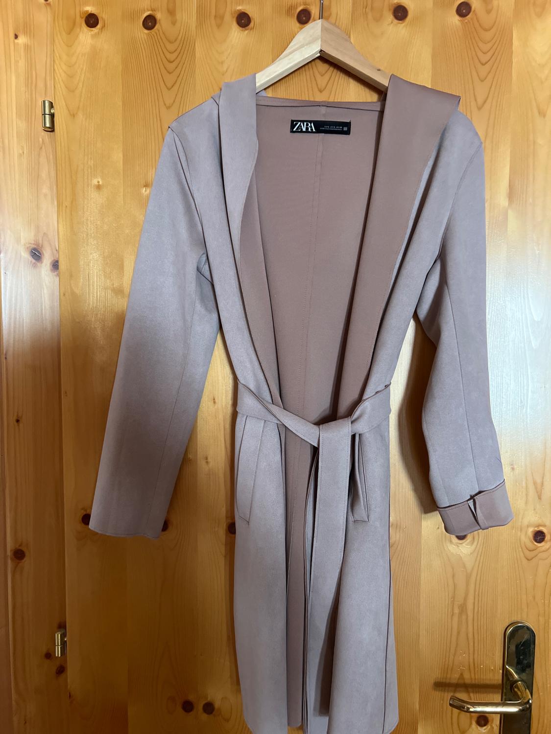 Light pink long coat with hood and belt, hanging on a hanger on a wooden door. Casual style. Visible text: ZARA.