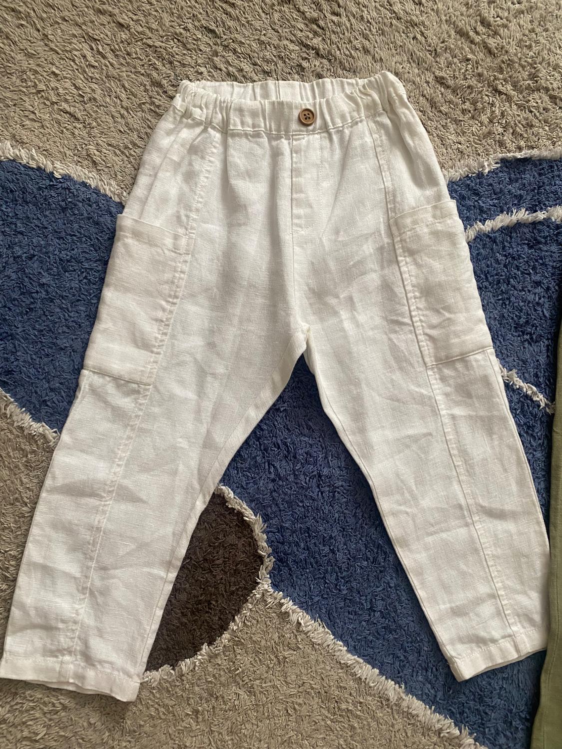 White linen trousers with elasticated waistband and wooden button, on a beige, blue and brown toned rug.
