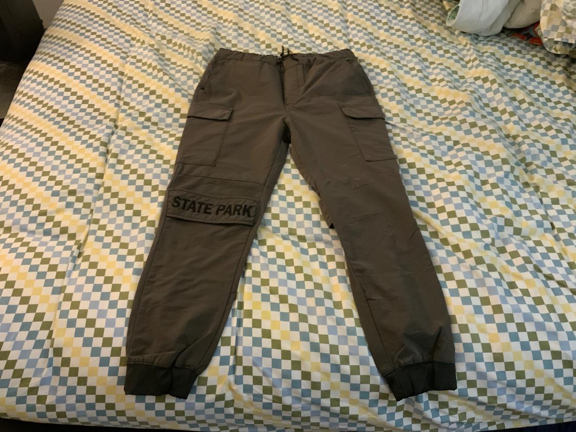 Olive green cargo jogger pants on patterned bed sheets. They have side pockets and the text "STATE PARK" on the leg.