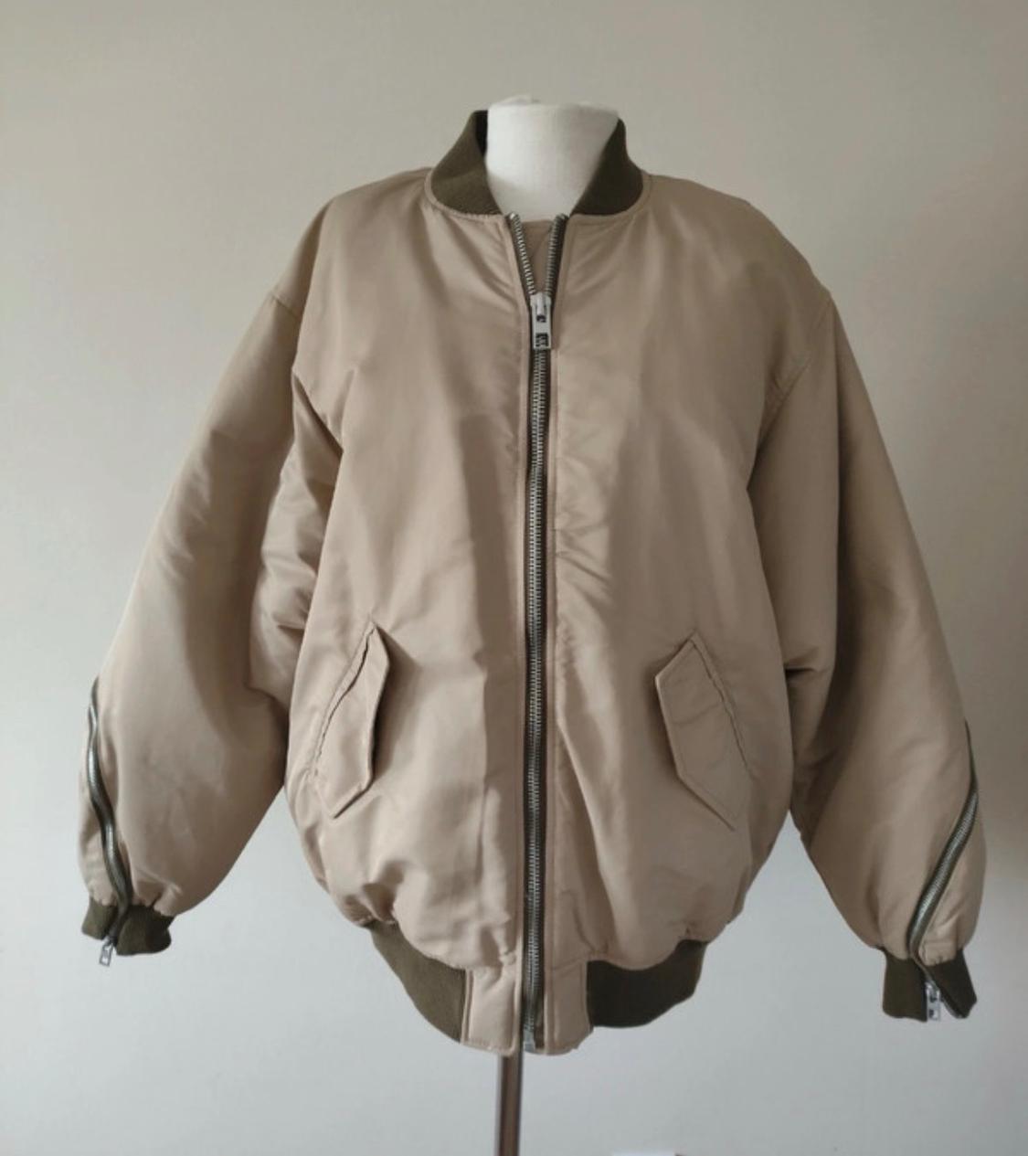 Beige bomber jacket on a mannequin. Design featuring a front zip, flap pockets, zips on the sleeves and olive-green edging.