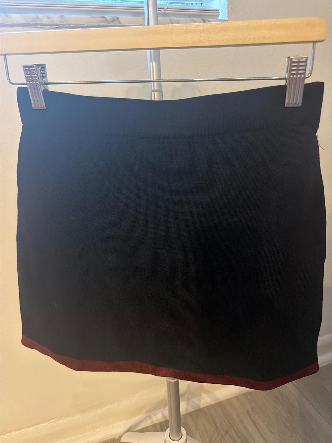 Short black skirt with a burgundy trim at the hem, hanging on a wooden hanger with clips on a metal rack in front of a white wall.