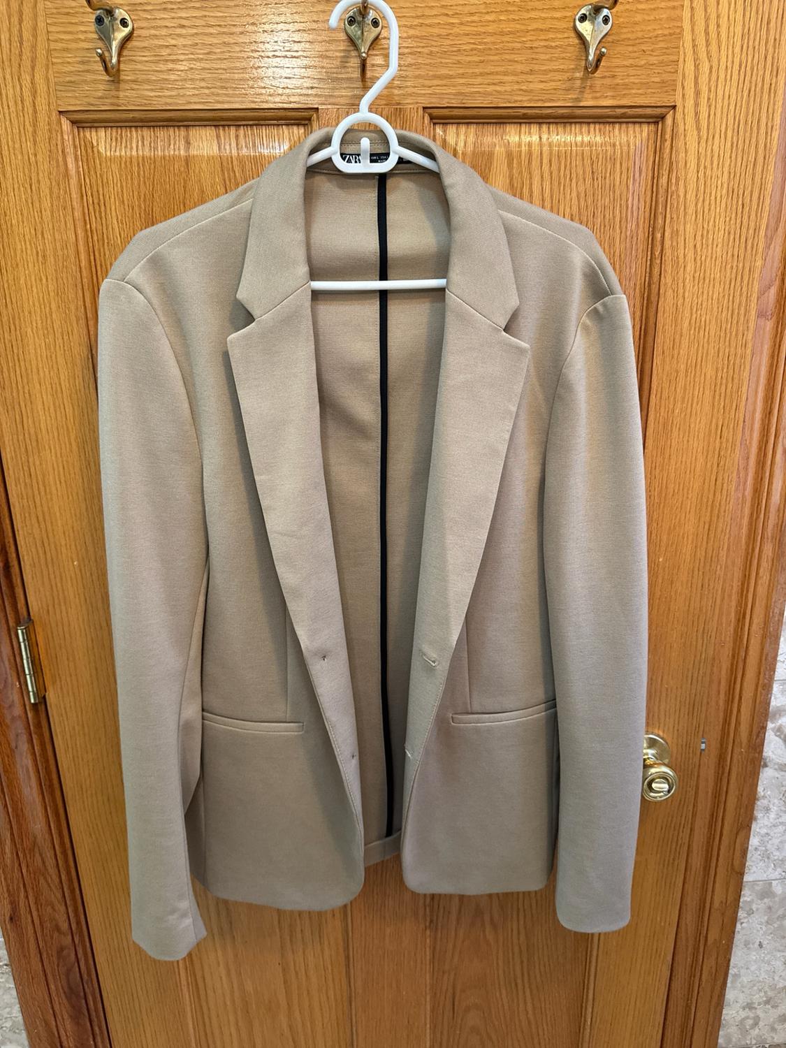 Beige blazer hanging on a white hanger against a wooden door. The interior label reads ZARA.