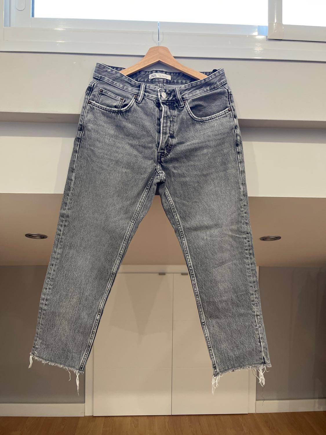 Distressed grey jeans hanging on a wooden hanger in front of a white wall. Frayed hem visible.