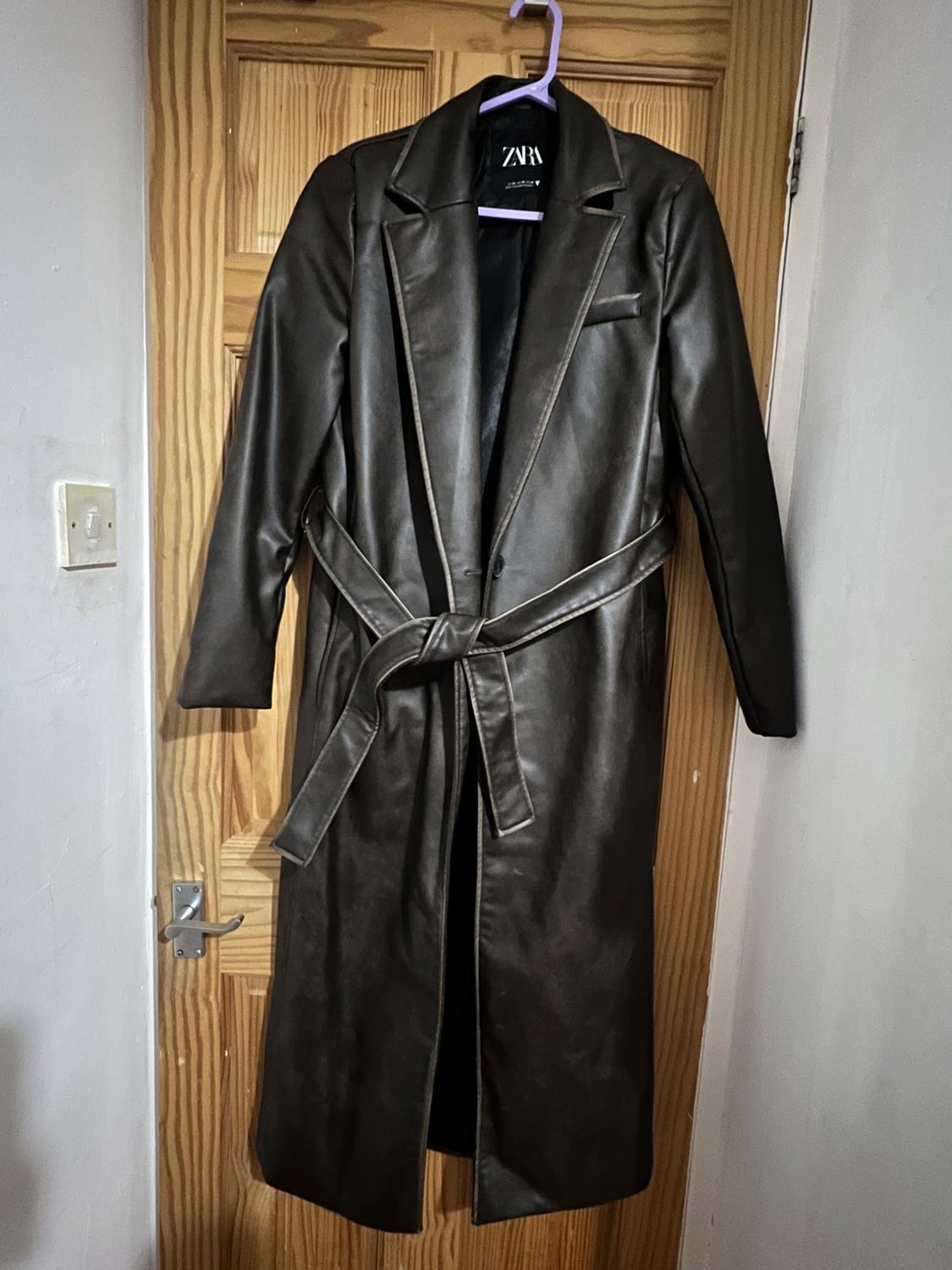 Long black coat hanging on a wooden door, with a belt and wide lapels. Visible label with the text "ZARA".