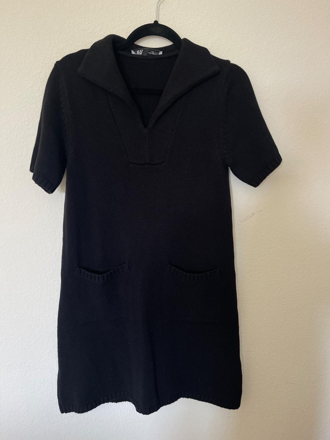 Black knitted dress with a wide neck and short sleeves, hanging on a black hanger against a white wall.