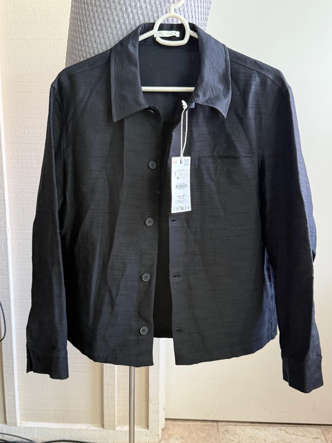 Black long-sleeved shirt with buttons and a chest pocket, hanging on a white hanger with a tag against a light background.