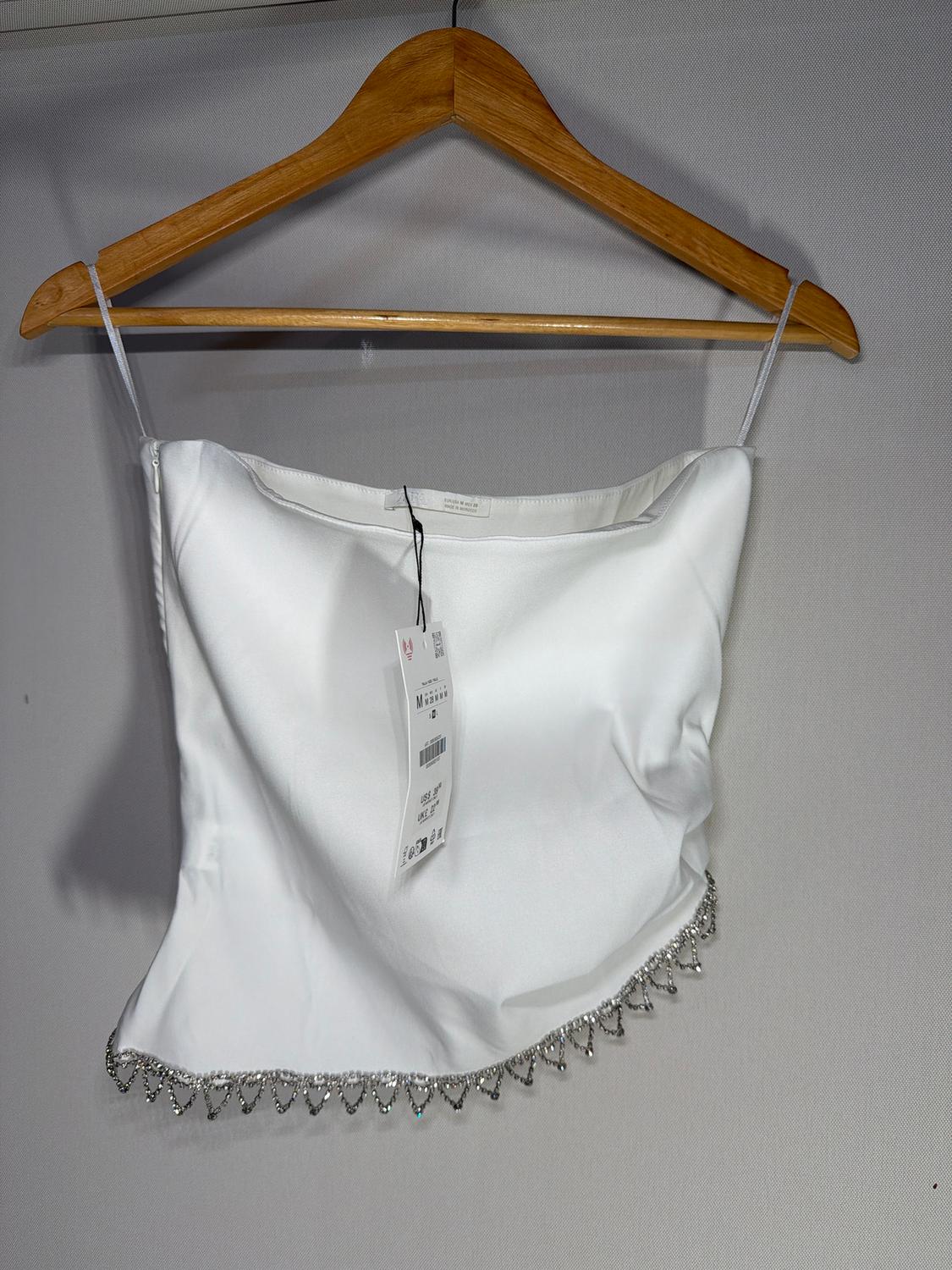 White camisole top with a diamante trim, hanging on a wooden hanger. Product details visible on the label.