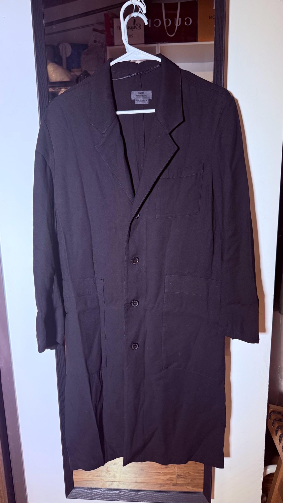 Long black coat hanging on a white hanger in front of a mirror. Lapel collar design, button fastening, and patch pockets.